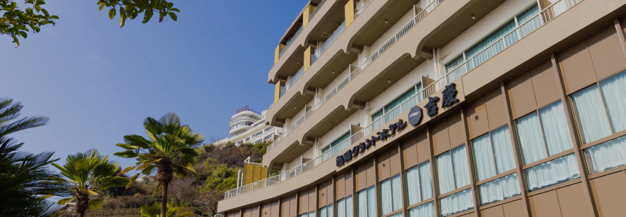 晴れ渡る青空を背景に建つ西浦グランドホテル吉慶の外観。ヤシの木がリゾート感を演出。