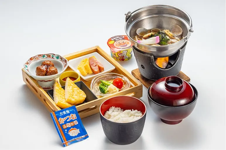 木製のトレイに色とりどりのおかずが並んだ、本格的な和食御膳。ミートボールや卵焼き、ウインナーに加え、火にかけた小鍋で温めるうどん、白ご飯、お味噌汁、デザートのムースがセットになっています。