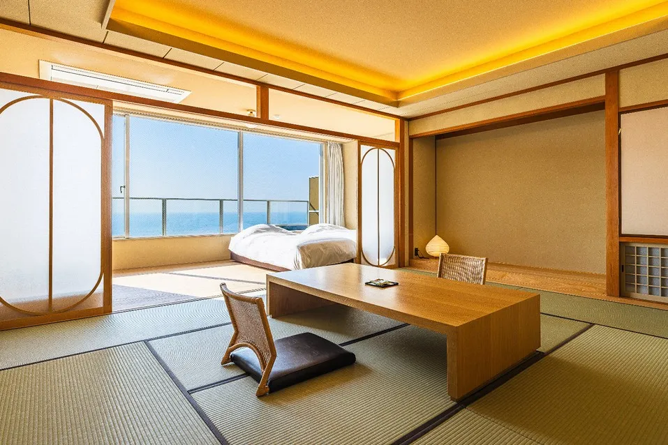 明るい光が差し込む、広々とした畳敷きの和室。手前には座椅子と木の座卓が置かれ、奥の大きな窓からは青い海が一望できます。天井の間接照明が温かみを添える、モダンで清潔感あふれる空間です。