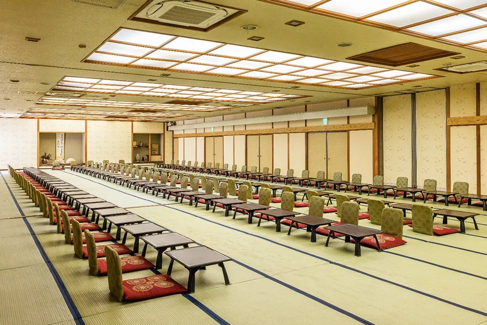 格天井（ごうてんじょう）のような美しい照明が印象的な、広大な畳敷きの宴会場。整然と並んだ一人一膳の座卓と座椅子が、奥まで真っ直ぐに続いています。床の間や掛け軸が飾られた伝統的な和の空間で、大人数のグループやご親族での集まりでも、皆さまで顔を合わせながら賑やかにお食事をお楽しみいただけます。