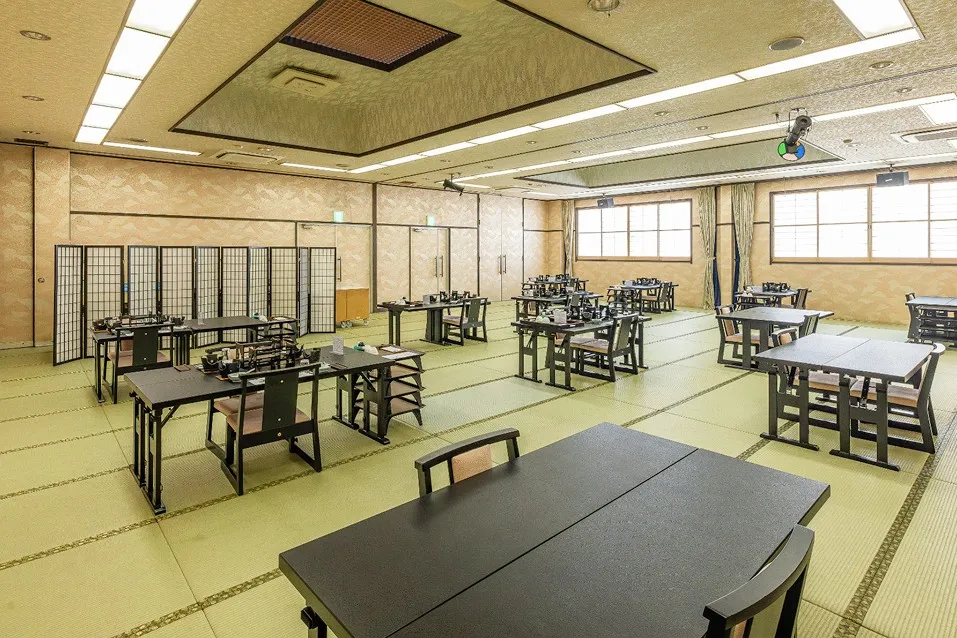 畳敷きの広間に、黒いテーブルと背もたれ付きの椅子がゆったりと配置された宴会場。和の趣を感じながらも、足への負担が少ないスタイルでお食事を楽しめます。衝立（ついたて）による仕切りや、外光が入る明るい窓際など、グループごとに落ち着いて過ごせるよう配慮された、モダンな和の空間です。
