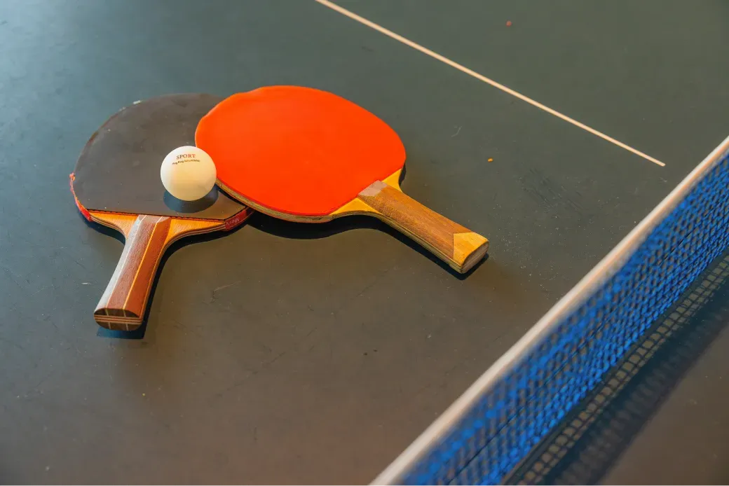 清潔感のある卓球台の上に置かれた、赤と黒のラケットと白いピンポン玉。