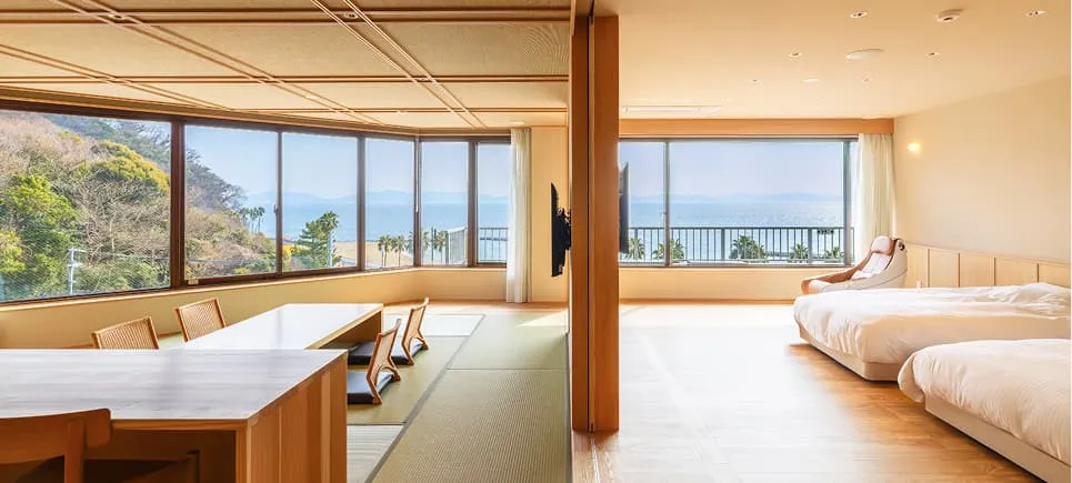 リニューアルされた特別室の全景。段差のないフラットなフロアに、左手には座卓のある畳スペース、右手にはツインベッドが配置されています。正面の大きな連続窓からは、三河湾の青い海と緑豊かな景色がパノラマ状に広がっています。