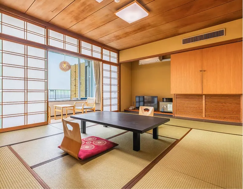 畳敷きの和室。中央には黒い座卓と座椅子、奥には海を望む広縁（ひろえん）があります。障子を開けると、大きな窓の向こうに青い海が広がり、穏やかな光が室内を照らしています。