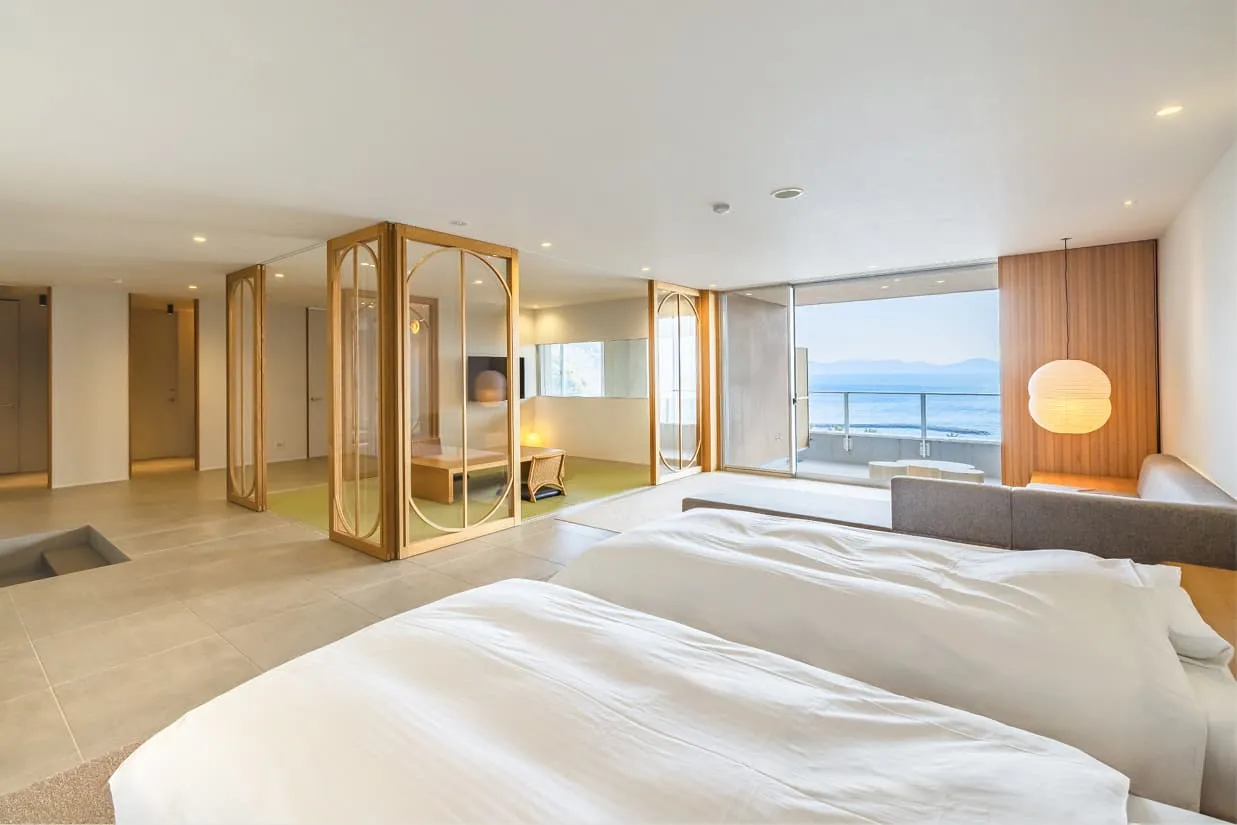 白を基調としたモダンな和洋室。手前には清潔感のあるデュベスタイルのベッドが並び、奥にはガラスの仕切りで囲まれた畳敷きの和室スペースがあります。大きな窓の外にはバルコニーと青い海が広がり、室内には柔らかなデザインの照明が灯る、上質な寛ぎの空間です。