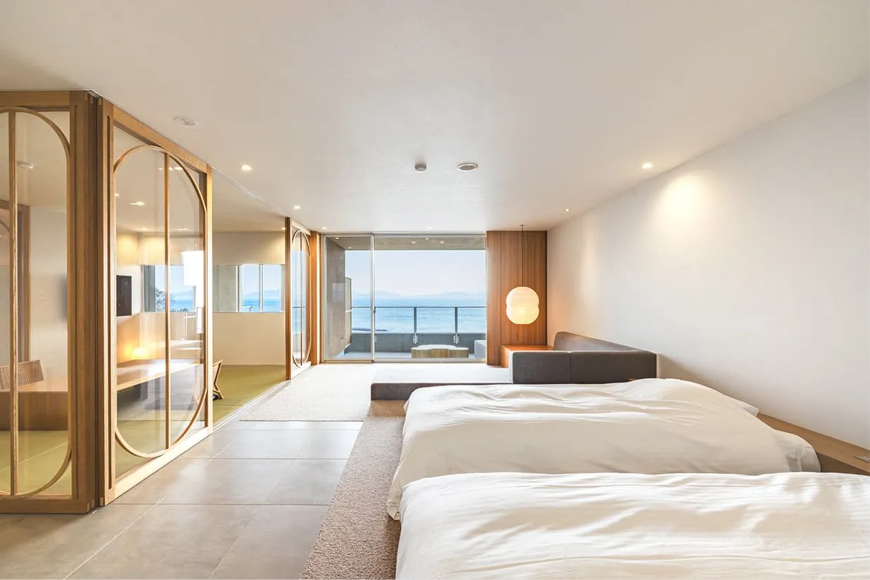 清潔感のある白を基調とした、モダンな和洋室。手前には心地よさそうなベッドが並び、ガラスで仕切られた畳のスペースには和風の照明が温かく灯っています。