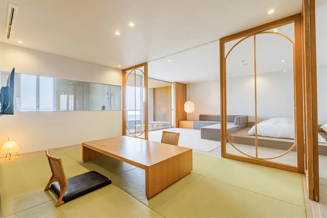 客室『wan~湾~』の和室スペース。手前には畳に木目の座卓と座椅子があり、ガラスの引き戸越しに奥のベッドルームやリビング、そして海へと視線が抜ける開放的な造りです。