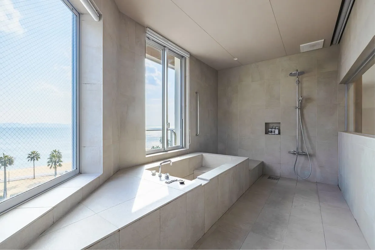 落ち着いたグレーの石造りの浴室に、スクエア型の広々とした浴槽が設えられています。大きな窓からは、青い海と砂浜、ヤシの木が並ぶ南国のような風景を眺めることができ、心地よい光が差し込む贅沢なバスルームです。