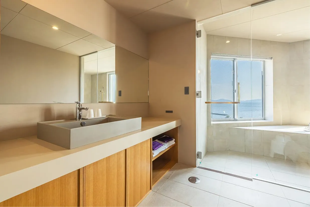 客室『wan〜湾〜』のパウダールーム。手前には横に長い広々とした洗面カウンターと大きな鏡があり、その奥には透明なガラスの扉を隔てて、海が見える半露天風呂へと繋がっています。全体がベージュや木目調の柔らかな色調で統一され、窓からの自然光が差し込む明るく開放的な造りです。