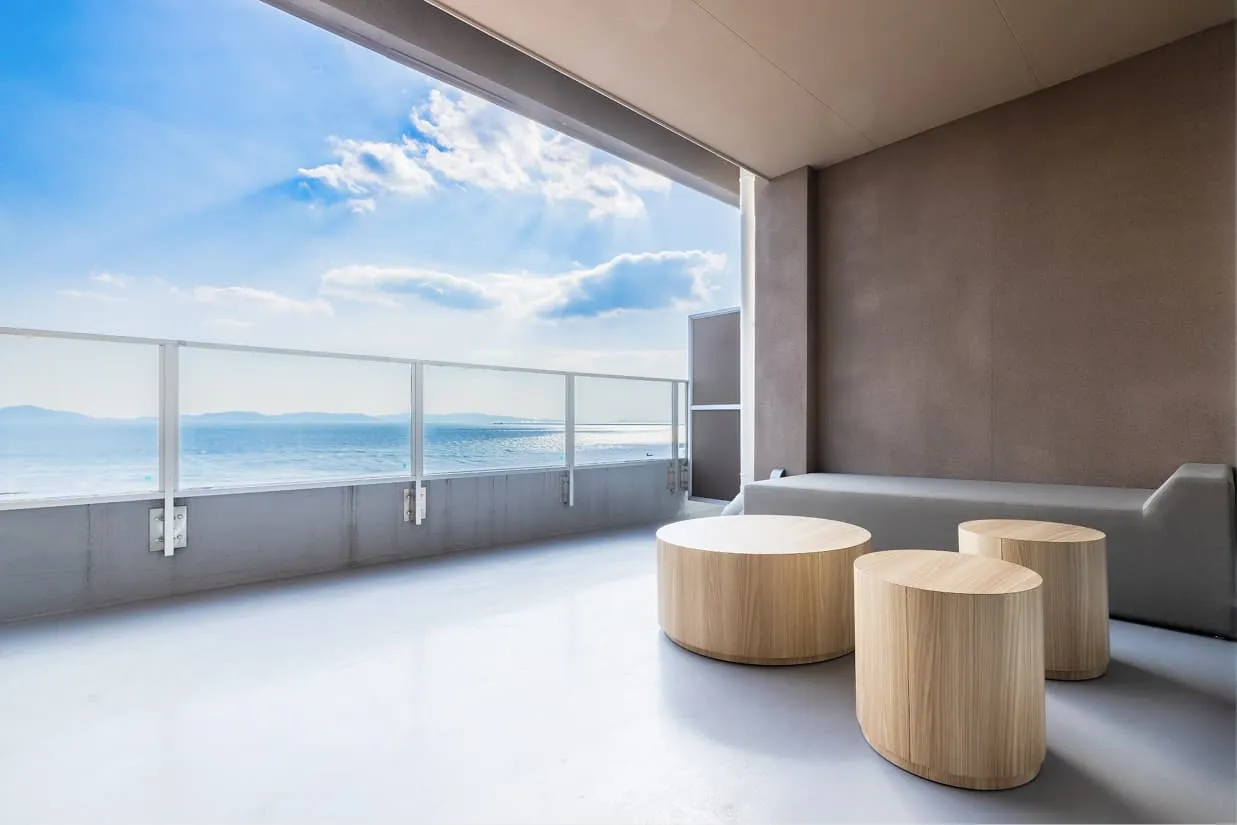 客室『wan〜湾〜』の広々としたバルコニー。木目の丸いテーブルとソファベンチが置かれたテラス席からは、遮るもののない大パノラマの海と空が広がります。