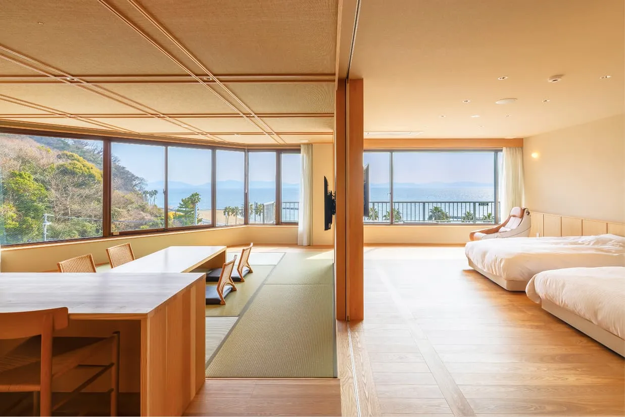 明るい日差しが差し込む「和洋室」の特別室。左側には畳敷きの和室とダイニングテーブル、右側には2台のベッドが並ぶ洋室があり、中央の引き戸で仕切られています。正面の大きな窓からは青い海と海岸線が一望でき、車椅子でも移動しやすそうな広々としたフラットな造りです。