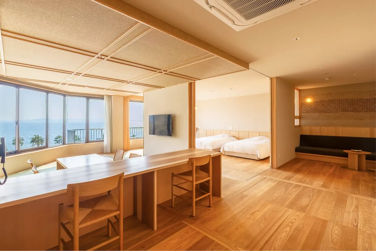 手前から奥へと続く広々としたユニバーサルデザインの客室。左側には海が見える畳スペース、中央には2脚の椅子が並ぶ長い木製カウンター、奥にはベッドルーム、そして右側の壁沿いには落ち着いたソファコーナーがあります。