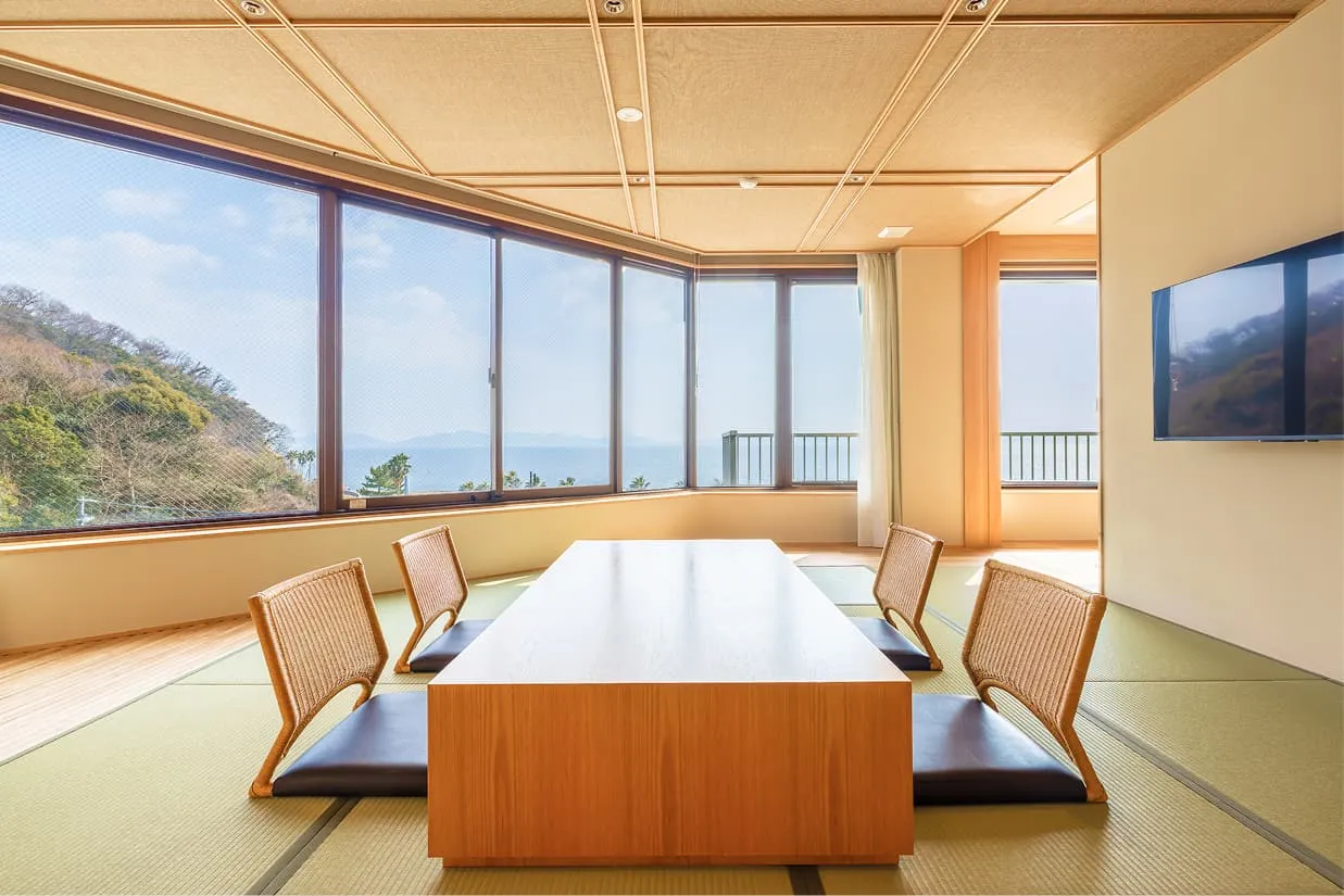 窓一面に広がるオーシャンビューを目の前に、畳敷きの和室スペースを中央から捉えた写真。大きな木製テーブルを囲むように、背もたれのある座椅子が4脚並んでいます。