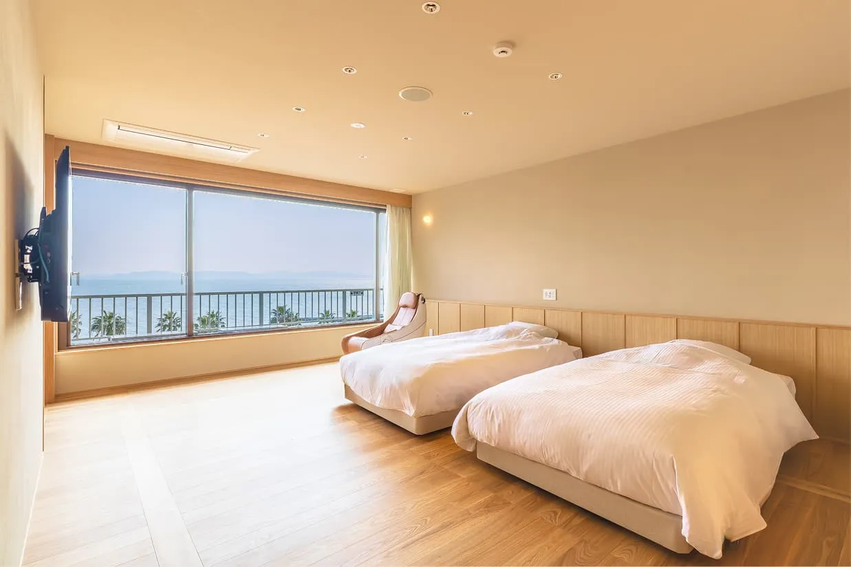 洋室の寝室エリア。右側の壁に沿って、清潔感のある白いリネンで整えられた2台のシングルベッドが並んでいます。ベッドの足元から左側の窓にかけて、車椅子でも余裕を持って回転できるほど広々としたフローリングのスペースが確保されています。