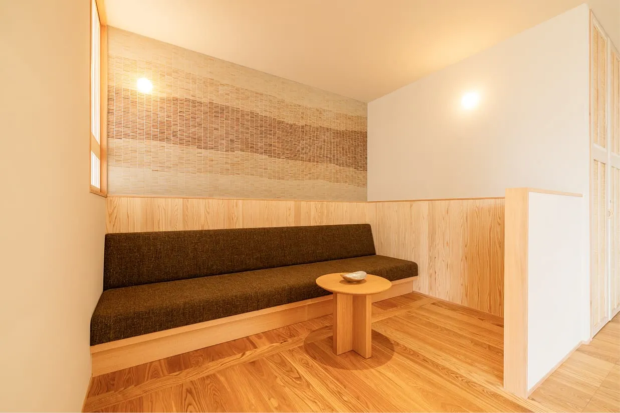 客室の一角にある、落ち着いた雰囲気のソファコーナー。壁のくぼみに収まるように設置された長いローソファと、小さな円形の木製サイドテーブルが置かれています。ソファ背面の壁は、地層をイメージしたような温かみのあるボーダー柄のタイルで装飾され、柔らかな間接照明がリラックスタイムを演出しています。