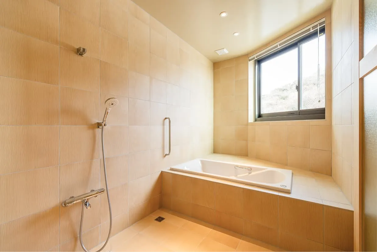 明るいベージュのタイルに包まれた、広々としたバスルーム。奥には、座った状態からでも移乗しやすいよう、周囲にゆとりあるスペースが確保された白い浴槽があります。浴槽横の壁には縦型の手すりが設置されており、立ち座りをサポートします。