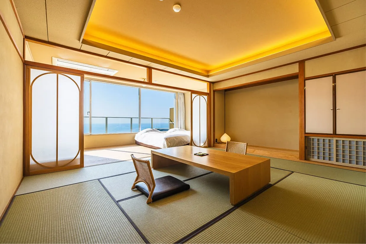穏やかな海を一望できる和洋室の客室。手前には畳敷きの和室に木製の座卓と座椅子があり、奥の広縁にはベッドが配置されています。天井の間接照明が温かみのある空間を演出し、大きな窓からは青い空と海が広がっています。