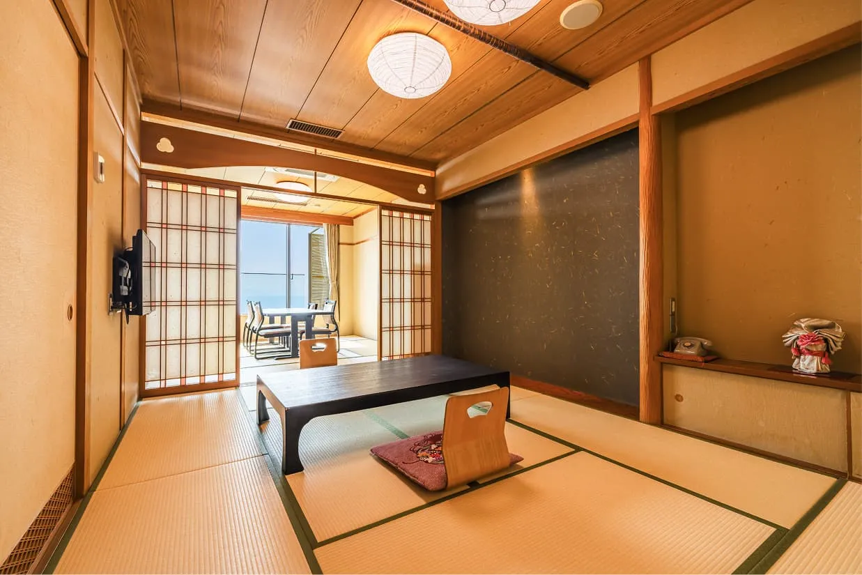 温かみのある木目天井と畳が心地よい、伝統的な和室。中央には座卓と座椅子があり、奥の障子を開けた先には、青い海を一望できる広々としたテラスとダイニングセットが見えます。壁の一部にはシックな濃紺のアクセントウォールが施され、和モダンな落ち着いた雰囲気です。