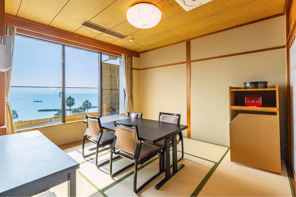 大きな窓から青い海とヤシの木を望む、開放感あふれるダイニングスペース。畳の上にモダンな黒のダイニングセットが置かれ、景色を眺めながら食事ができる造りです。