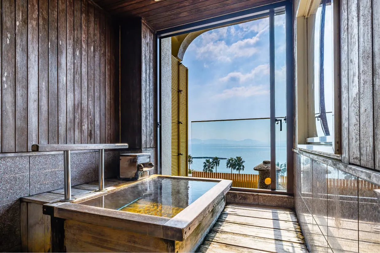 客室専用の木製露天風呂。使い込まれた木の質感が心地よい湯船には、なみなみとお湯が湛えられています。目の前の大きな開口部からは、穏やかな青い海と空、そして海岸沿いのヤシの木を一望でき、波音を感じながら贅沢な湯浴みを楽しめるプライベート空間です。