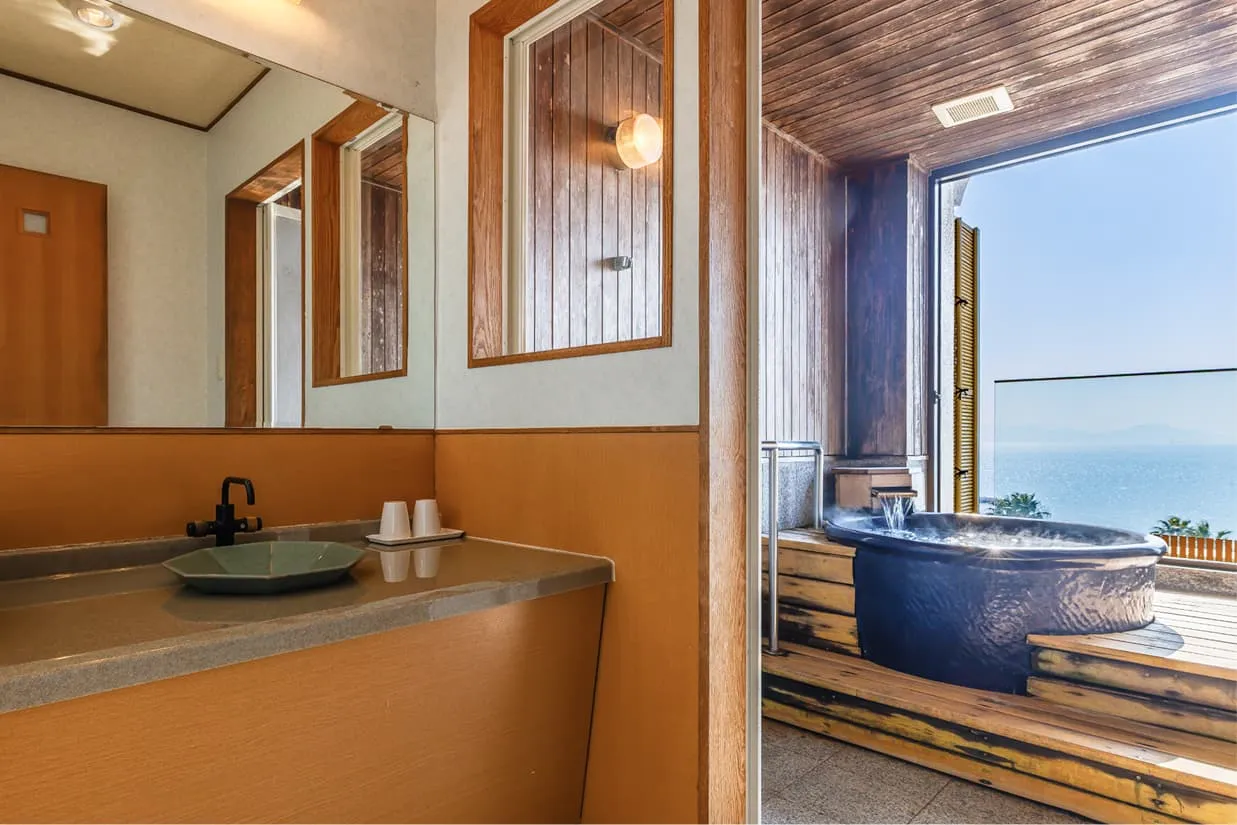 洗面スペースから露天風呂へと続く開放的な眺め。手前には、和の趣がある落ち着いた色調の洗面台があり、その右隣には、海に面した客室専用の露天風呂が広がっています。