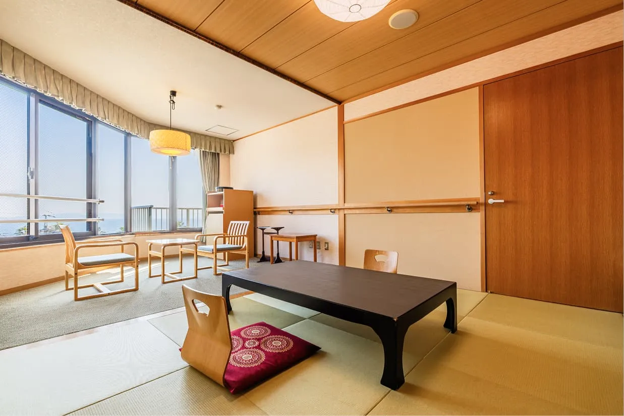 畳が敷かれた和室スペースと、海を望む広縁（ひろえん）。壁面には伝い歩きをサポートする木製の手すりが設置されています。中央には座卓と背もたれ付きの座椅子があり、奥の広縁には立ち上がりやすい高さの肘掛け付き椅子2脚とサイドテーブルが置かれています。