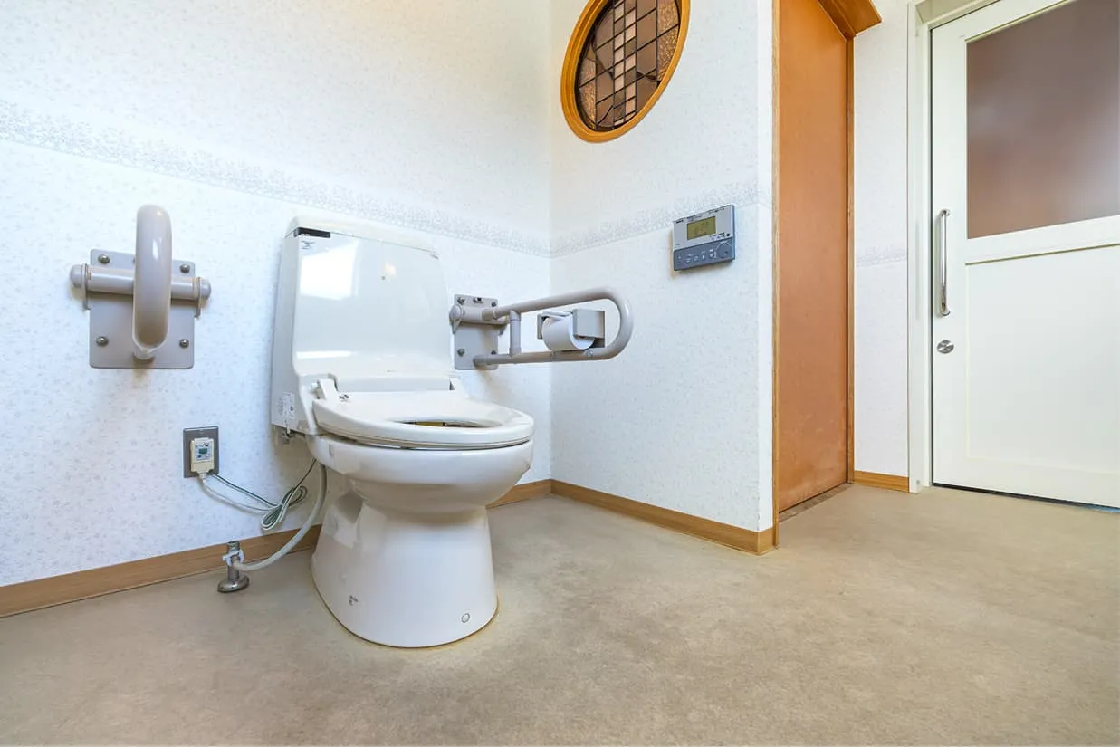 車椅子でもゆったりと回転できる広さを確保したバリアフリー設計のトイレ。便座の両側には手すりが設置されており、向かって右側は跳ね上げ式、左側は縦型の手すりになっています。入り口は段差のない引き戸で、白を基調とした清潔感のある空間です。