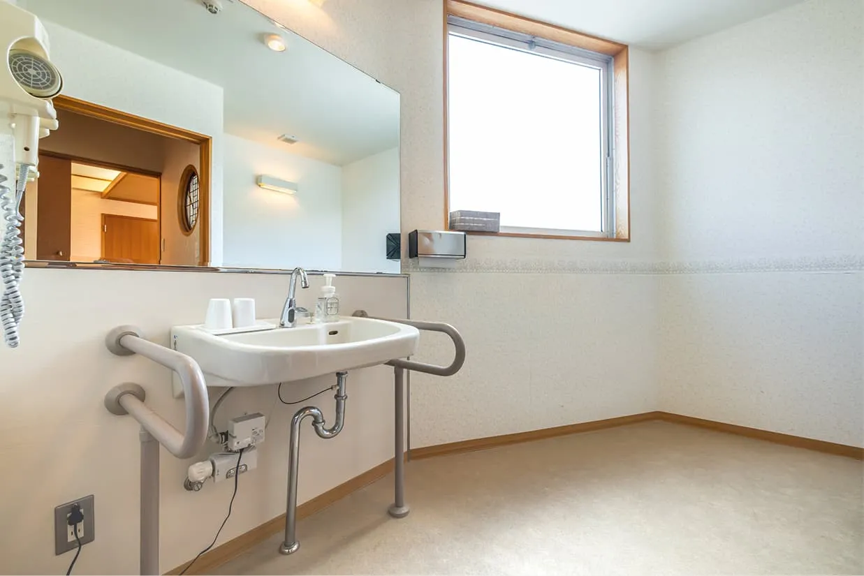 車椅子対応の広々とした洗面スペース。洗面台の足元はオープンになっており、車椅子のまま奥まで入り込める設計です。洗面ボウルを囲むように頑丈な手すりが設置されており、立ち上がりの補助や姿勢の保持に役立ちます。