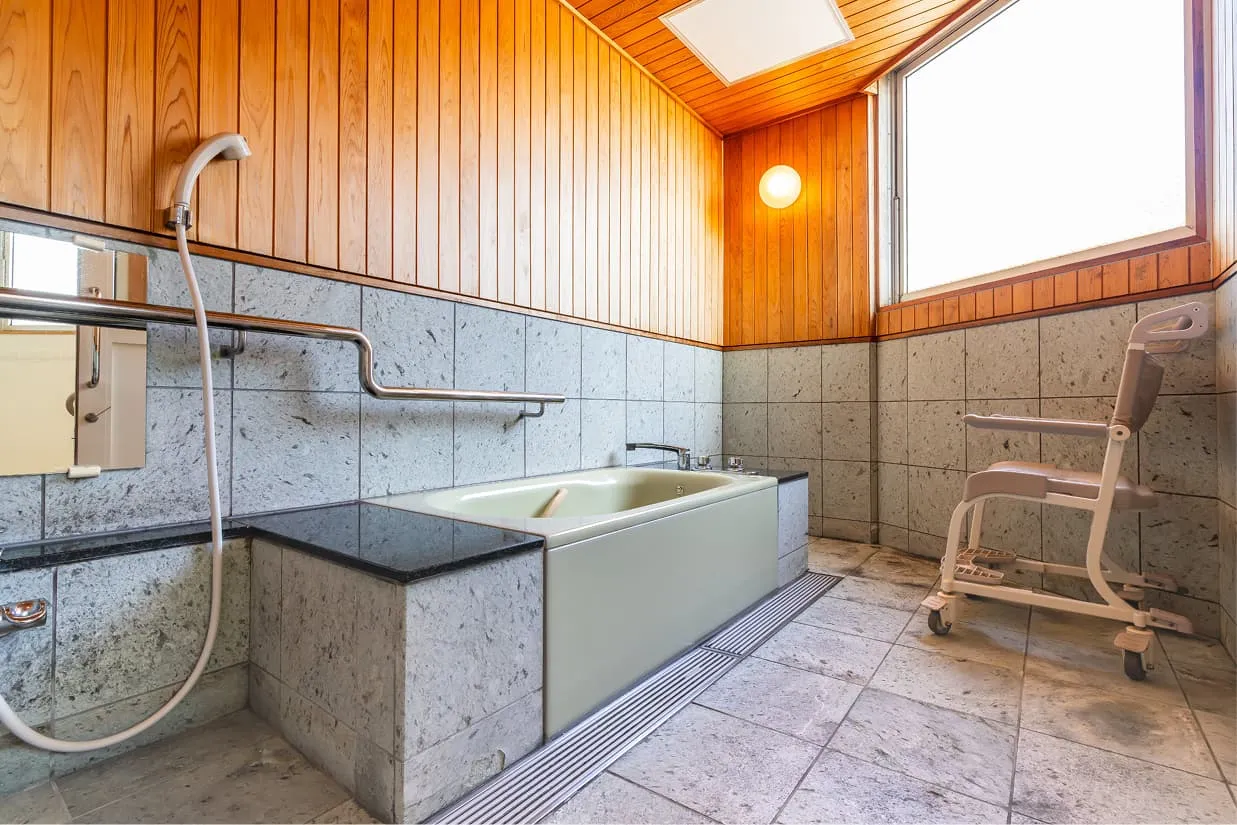 石造りの壁と木の天井が美しい、広々としたバリアフリー浴室。浴槽の横には、腰を掛けてから入浴できる黒い石材の移乗スペース（ベンチ）があり、壁面には横長の手すりが設置されています。室内にはキャスター付きのシャワーチェアも用意されており、洗い場は介助者も一緒に動ける十分な広さがあります。