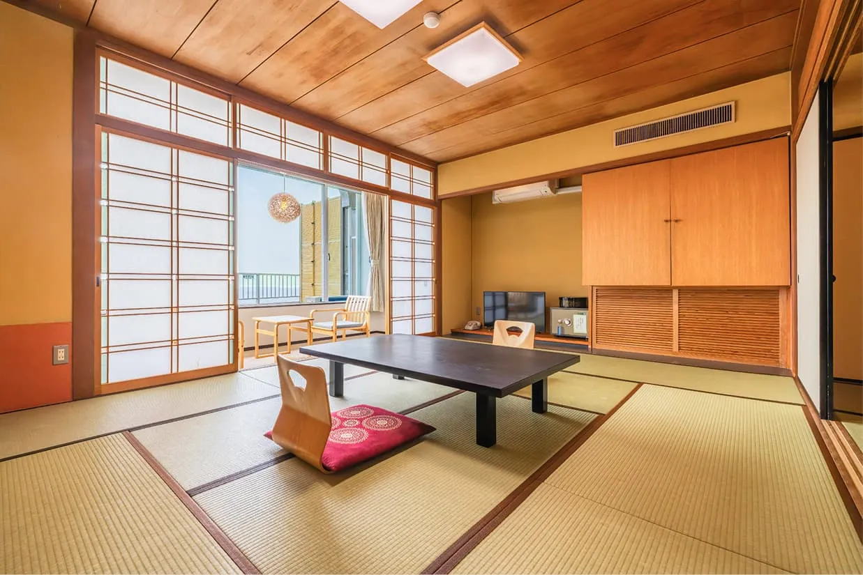畳が敷かれた伝統的な和室の客室。中央には黒い座卓と座椅子、赤い座布団があり、奥の大きな障子窓からは明るい海が見えるオーシャンビューの景色が広がっています。天井は木目調で、木のぬくもりを感じる落ち着いた雰囲気の空間です。