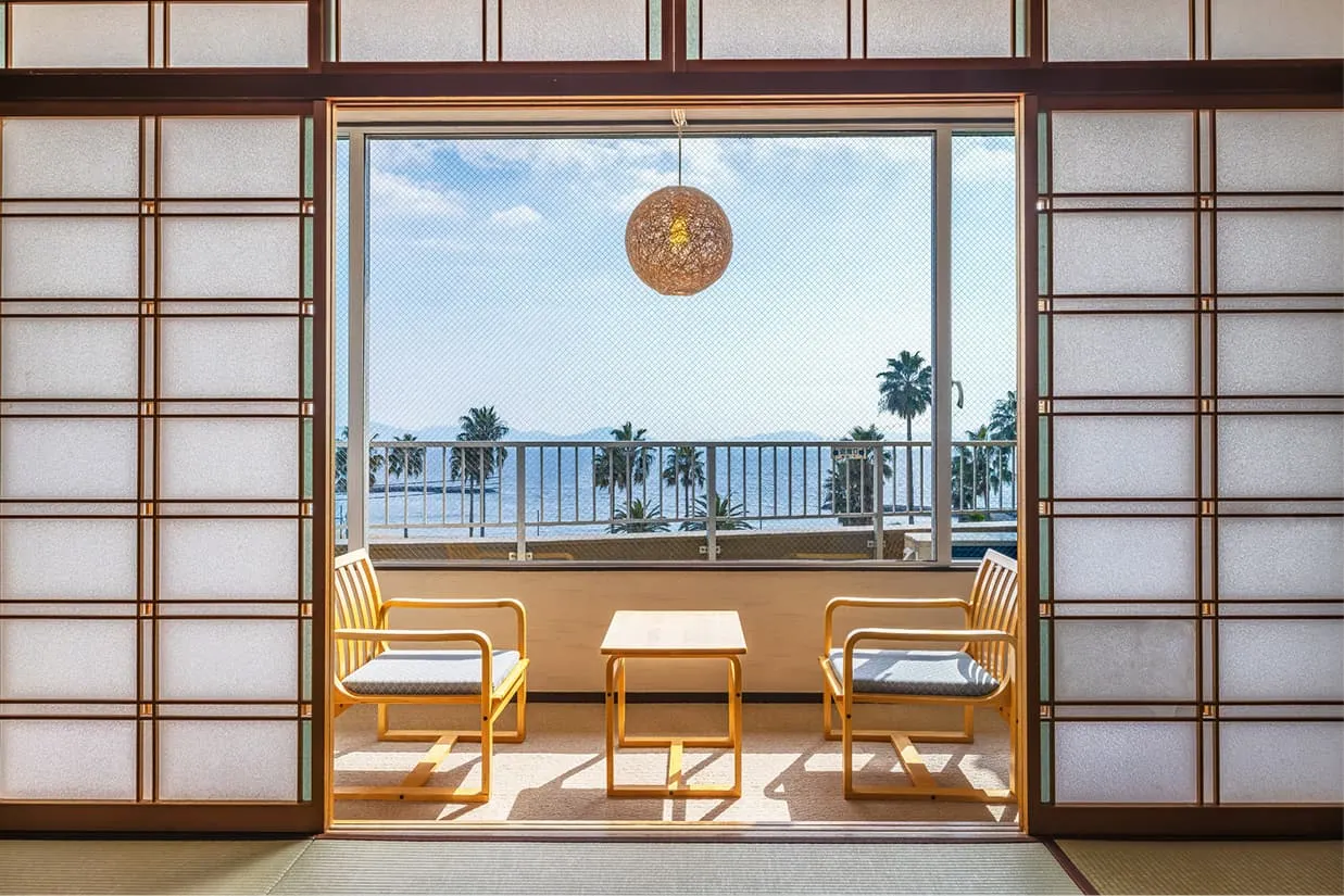 左右に開かれた障子の向こうに、青い海とパームツリーが広がる和室の広縁。窓際には木製の椅子2脚と小さなテーブルが置かれ、天井からは丸い麻玉の間接照明が吊り下げられています。明るい日差しが差し込む、開放感あふれる景色です。