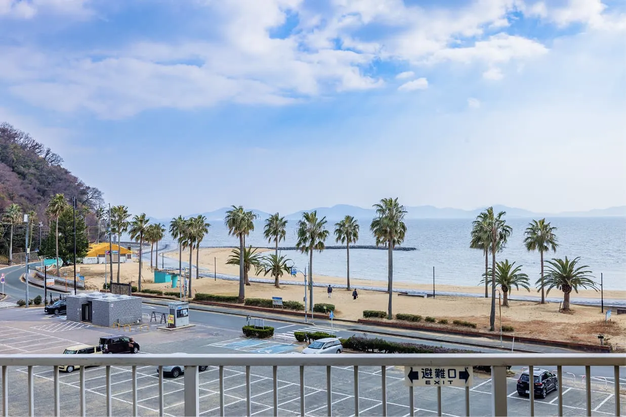 客室のベランダから見下ろすオーシャンビューの景色。手前には駐車場があり、その先に砂浜とヤシの木が並ぶ海岸線が広がっています。穏やかな青い海と遠くの山々、青空が広がる開放的な昼前の風景です。