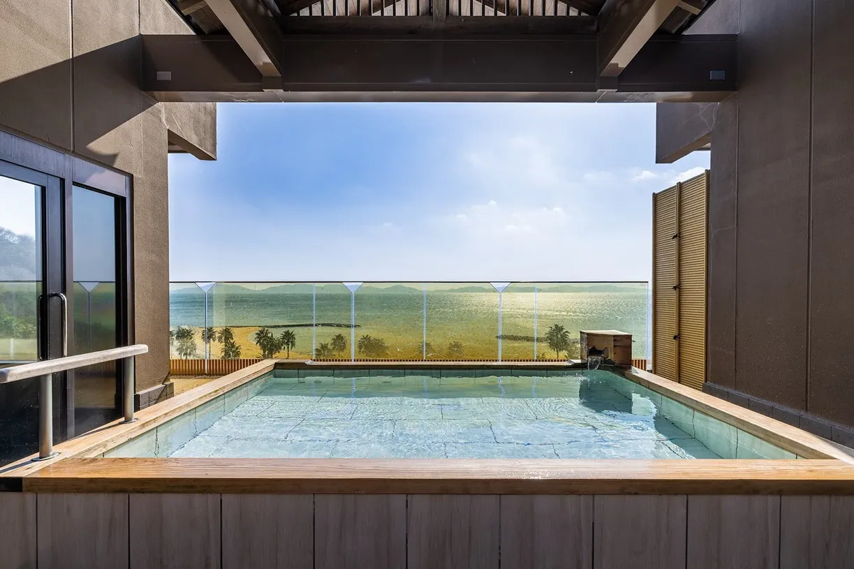 額縁のような開放的な柱の間から望む、展望露天風呂。手前には四角い木枠の湯船があり、その向こうには陽光を反射してきらめく三河湾の水平線と、青い空がどこまでも広がっています。