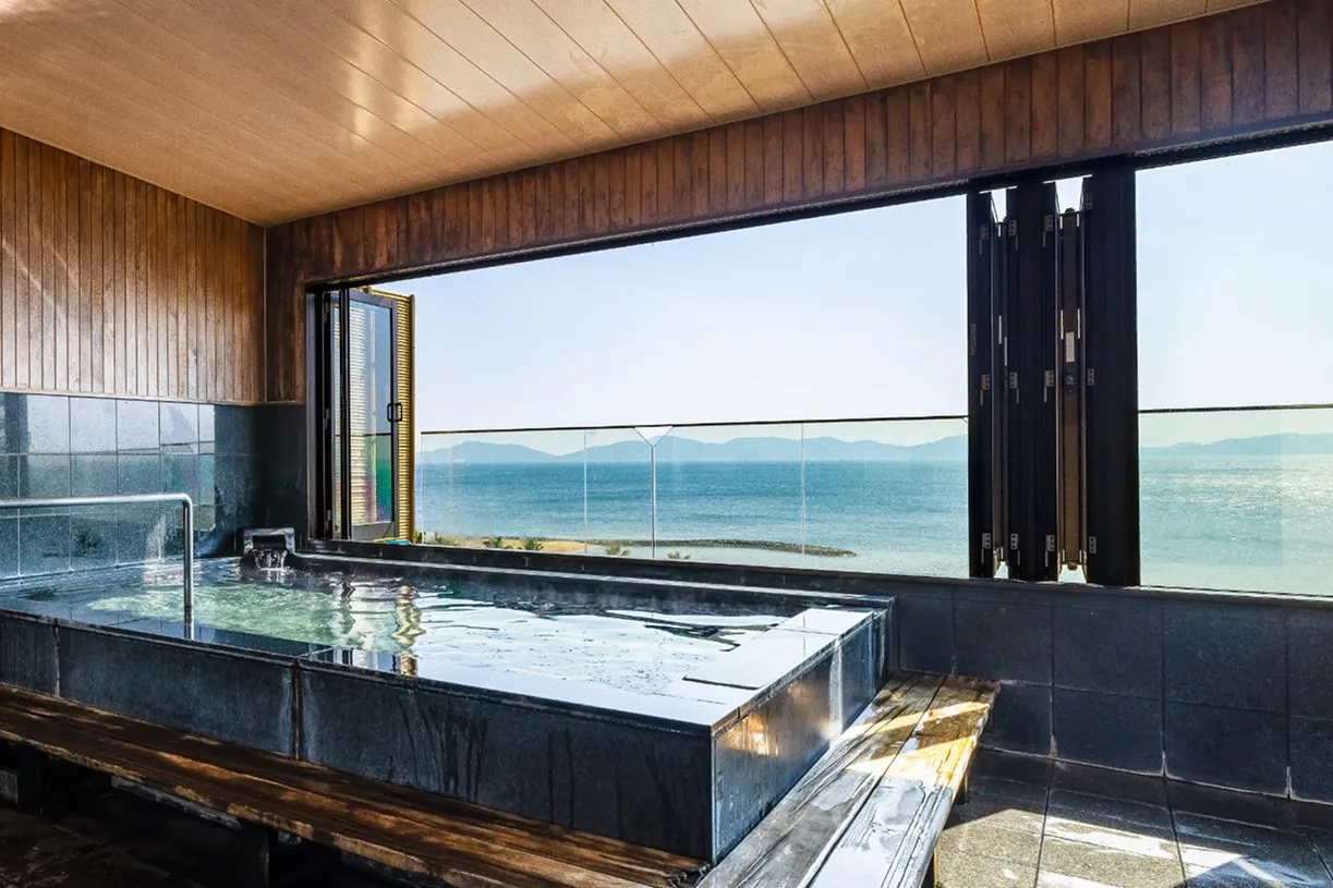 斜め方向から見た屋内展望風呂。手前から奥へと続く黒い石造りの湯船に、大きく開かれた窓から三河湾の明るい陽光が差し込んでいます。