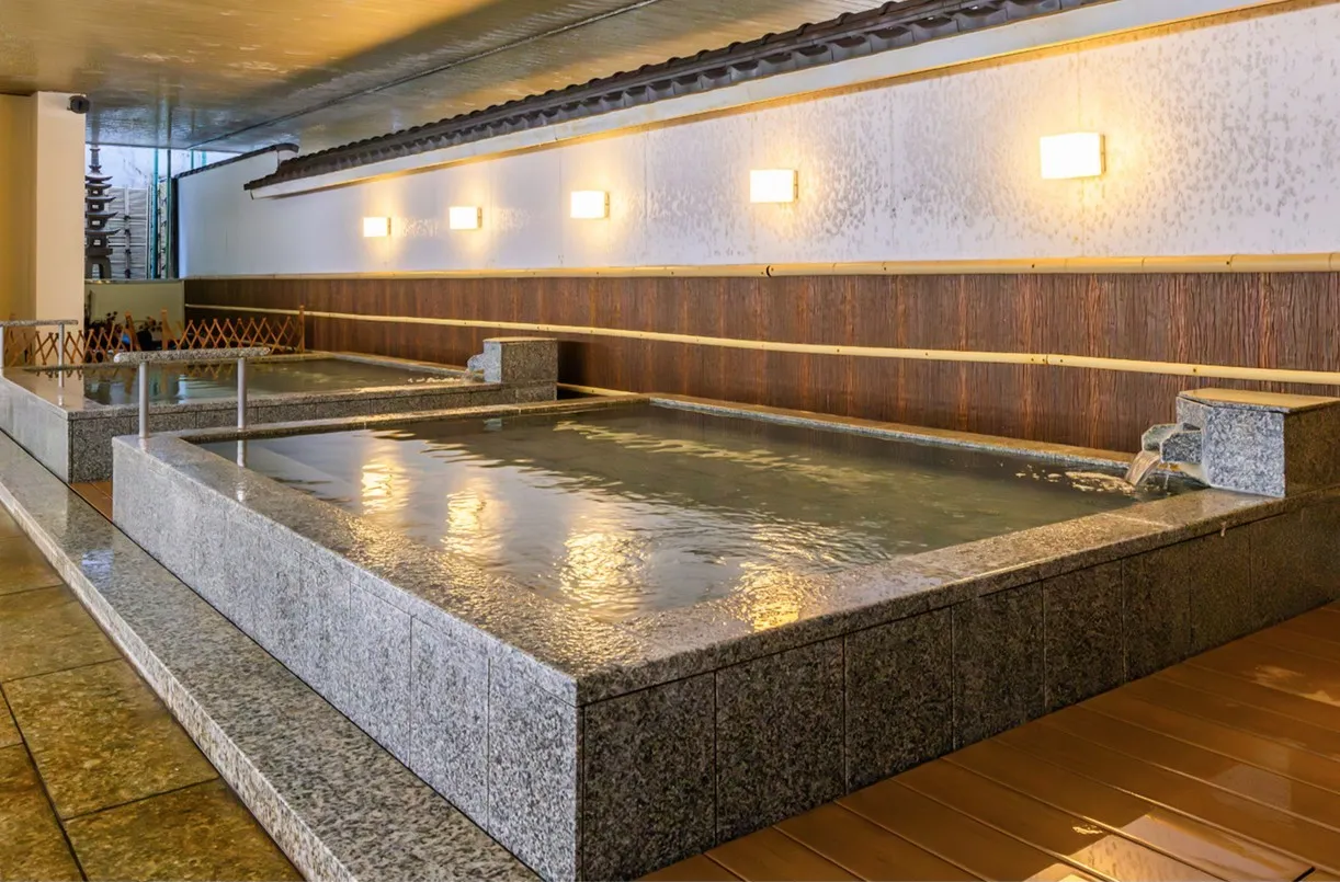 手前から奥へと二つの大きな石造りの浴槽が並ぶ、落ち着いた屋内大浴場。白と茶色の和モダンな壁面には四角い照明が等間隔に灯り、温かな光が静かな水面に美しく映り込んでいます。