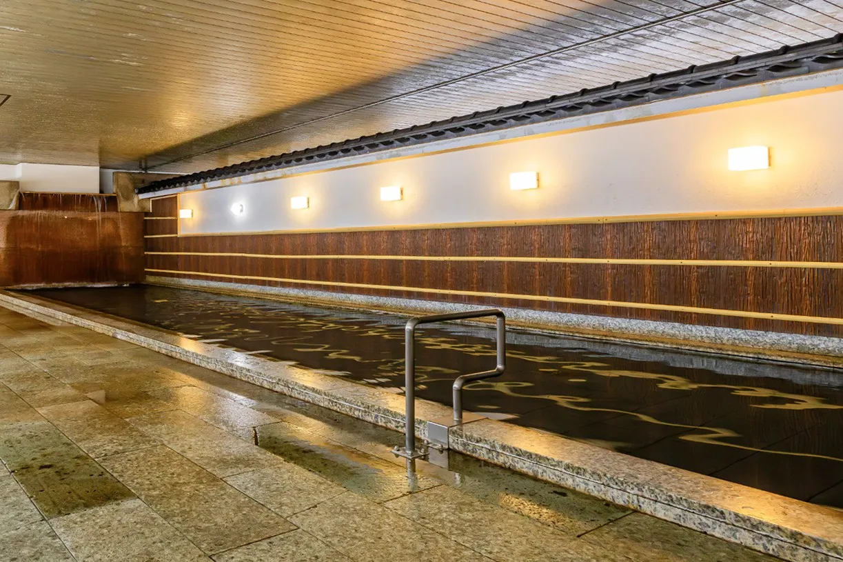 横に長く広がる石造りの大浴場。奥へと続く壁面には温かみのある四角い照明が並び、その光が静かな水面に幾筋もの波紋のように反射して、幻想的な和の空間を作り出しています。