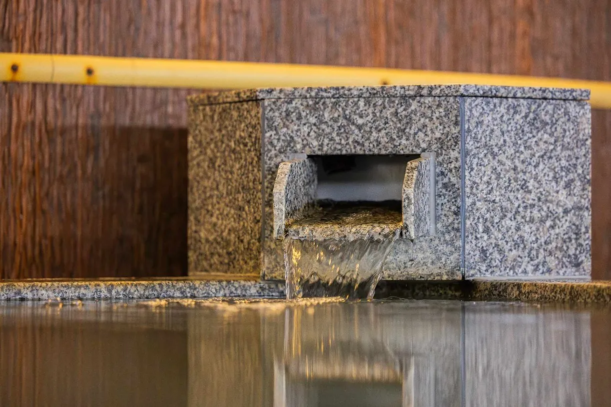 和風の壁際にある、御影石の湯口。さらさらと清らかな温泉が絶え間なく注がれ、温かみのある照明を反射する静かな水面へと溶け込んでいく、趣深い情景。