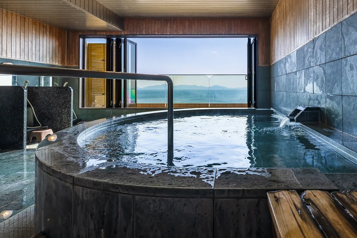 手前にゆったりとした曲線を描く石造りの湯船。大きく開かれた正面の窓からは、光り輝く三河湾が一望でき、反射した光がお湯の表面をやさしく照らしています。