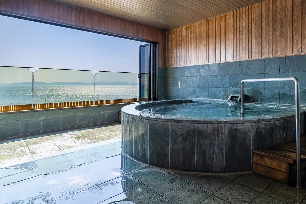 右側に円形の石造りの湯船を配した展望風呂。大きく開かれた窓から差し込む陽光が、濡れたタイル床に鏡のように反射してキラキラと輝いています。