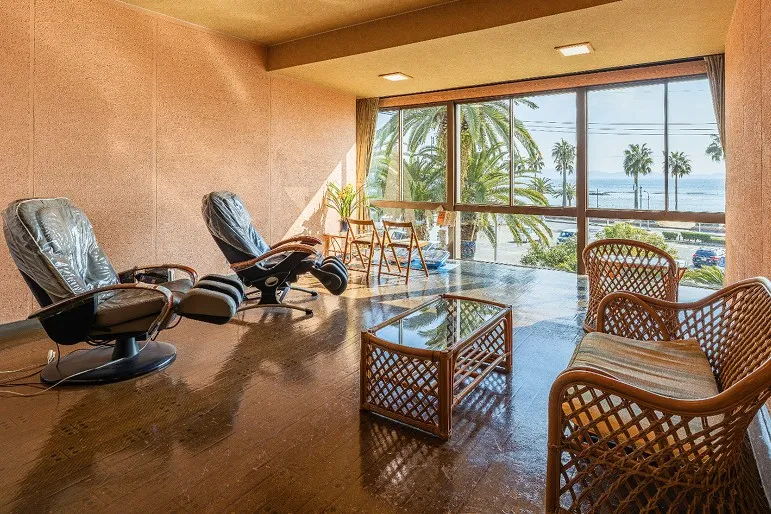 大きな窓の向こうに青い海とヤシの木が広がる、開放的なラウンジの写真です。手前にはゆったりとした藤編みのソファ、窓側には海を正面に臨む2台のマッサージチェアが並び、明るい陽光が差し込んでいます。